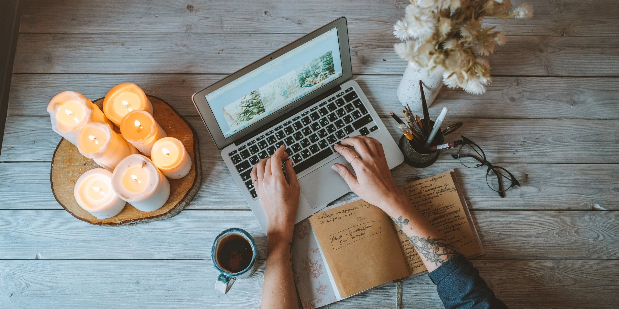 Birdseye image of women typing at a laptop on a desk, candles, notebook, coffee, glasses, pens and flowers in view